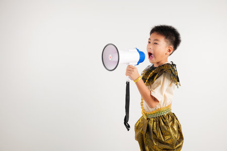 Portrait Thailand kid boy smiling traditional Thai dress costume shouting by megaphone, studio shot isolated white background, primary screeching through in megaphone announces discountsの写真素材
