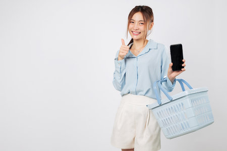 Smiling Asian woman with grocery basket and mobile phone shows thumb up, shopping experience in supermarket with phone screen display and online shopping cheer. isolated studio white backgroundの写真素材