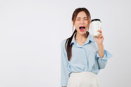 Sleep deprived Asian woman yawns while holding takeaway coffee paper cup, showing sleep exhaustion and tired falling asleep emotion in early morning routine with stress and mouth wide openの写真素材