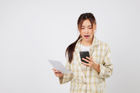 Shocked woman holding phone and invoice struggles with financial worries and financial stress while reviewing bill payments. isolated studio white backgroundの写真素材