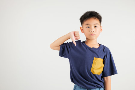 Portrait disheartened Asian young kid boy displaying a thumbs down gesture, symbolizing dislike. Studio shot isolated on white with ample copy space, primary child portraying negative and sad lookの写真素材