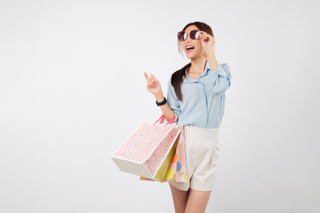 Excited asian young woman smile hands hold bags and glasses during Black Friday, touching her sunglasses, looking amazed and happy after a great shopping experience, isolated studio white backgroundの写真素材