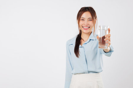 Happy Asian woman smile, young lifestyle care hold glass of water, a beautiful portrait of a model keeping healthy with daily water hydration routine for wellbeing, isolated studio white backgroundの写真素材
