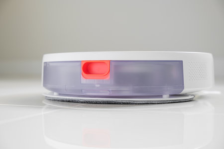 White automatic floor cleaner with side brush on tile flooring. Back view shows clear dustbin and red release button on white surface. Efficient smart home cleaning device for dust and debris removalの写真素材