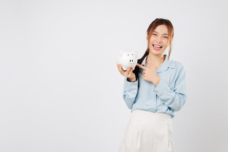 Portrait smile happy Asian woman holding piggy bank and pointing finger with piggy excitement saving money proudly and trusting her banking path for financial success. isolated studio white backgroundの写真素材