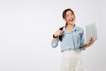 Happy young Asian woman smiling and holding a laptop showing thumbs up, feeling excited while working on another laptop computer for good business tasks. isolated studio white backgroundの写真素材