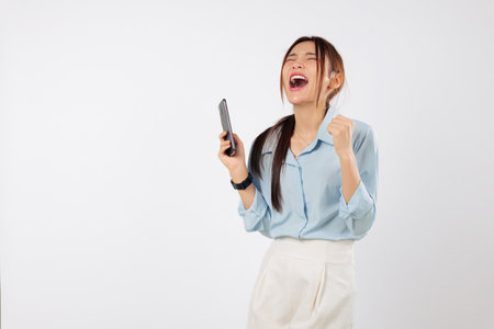 Happy young Asian woman holding a mobile phone with fists clenched, excitedly celebrating success. She is a winner, feeling the victory using her smart phone. isolated studio white backgroundの写真素材
