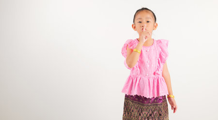 Portrait Thailand kid girl smiling traditional Thai dress costume making finger on lips mouth silent quiet gesture, studio isolated white background, kindergarten child marking silence for stop quietの写真素材