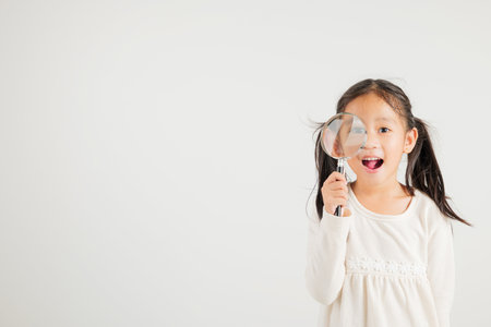 Asian little kid girl funny looking through magnifying glass at studio shot isolated on white background, Happy kindergarten child lifestyle smiling exploring holding magnifier searching, Educationの写真素材