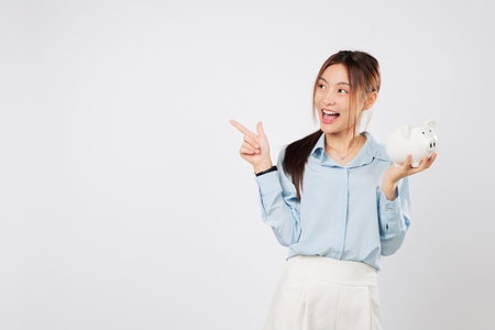 Portrait Happy Asian woman holding piggy bank while pointing with piggy excitement showing money saving plan and strong confidence in her banking steps. isolated studio white backgroundの写真素材