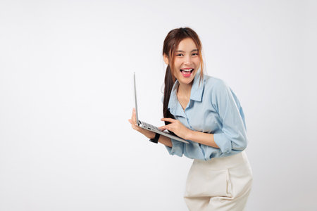 Laptop on desk as happy young Asian woman smiling types with energy, using another laptop screen nearby for dual-computer portable workflow and focus. isolated studio white backgroundの写真素材