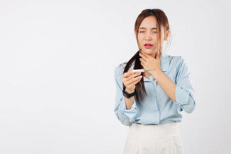Woman with flu has sore throat, checks temperature with thermometer, touching her neck feeling sick and unwell, having a high temperature, feeling sad as a young sick woman, isolated white backgroundの写真素材