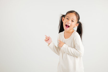 Happy Asian portrait cute young kid girl smiling indicate finger empty space, studio shot isolated on white background, Thai kindergarten child pointing index out with copy spaceの写真素材