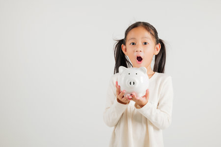 Asian little kid girl holding piggy bank and looking at camera at studio isolated on white background, Happy kindergarten child lifestyle smiling with is white piggybank, Thai personal money savingsの写真素材