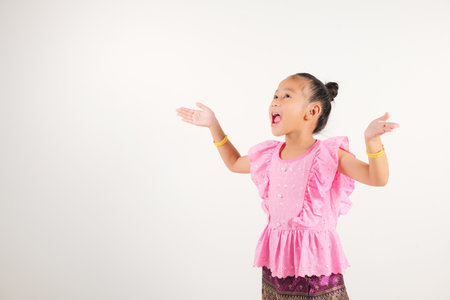 Portrait Thailand kid girl smiling traditional Thai dress costume raises fists in celebration of her success, studio isolated white background, kindergarten child saying yes with excitement winningの写真素材