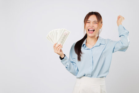 Asian woman smiles brightly holding money, raising her fist with excitement, celebrating lucky moment and extra money. hold currency raised fist isolated studio white backgroundの写真素材