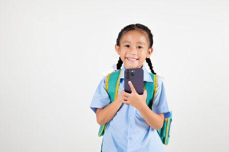 Portrait smiling Asian little girl kindergarten with backpack holding mobile phone studio isolated white background, happy woman kid in pigtails wearing school uniform use smartphone, back to schoolの写真素材