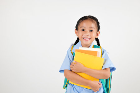 Portrait Asian smiling young girl kids kindergarten holding backpack and hugging books studio shot isolated white background, happy cute little schoolgirl wears school uniform, back to school conceptの写真素材