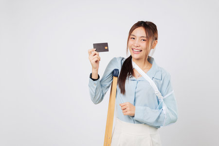 Smiling woman broken with crutches and splint arm shows credit card payment after insurance approval. Insurance helped her manage payment after injury and financial stress. isolated white backgroundの写真素材