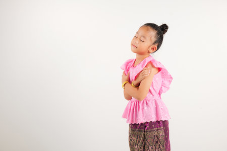 Portrait Thailand kid girl smiling traditional Thai dress costume hugging herself remember moment, studio isolated white background, kindergarten child love yourself carefree self care and self esteemの写真素材