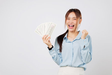 Happy woman cheers with raised fist holding money fan banknotes, celebrating lucky moment and feeling empowered by money and success. isolated studio white backgroundの写真素材