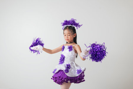 Smiling kid cheerleader in purple and white uniform, holding pom-poms. Studio shot with a clean...