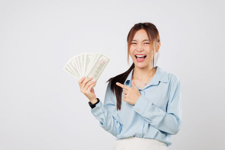 Happy Asian woman holding money fan banknotes and pointing to it with joy, showing dollar bills and celebrating money luck and winning. isolated studio white backgroundの写真素材
