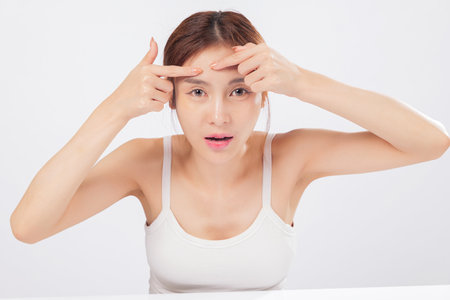 Acne removal in process as she targets acne on her forehead, this young woman handles skin imperfections with confidence despite the blemish and pimple worries. isolated studio white backgroundの写真素材