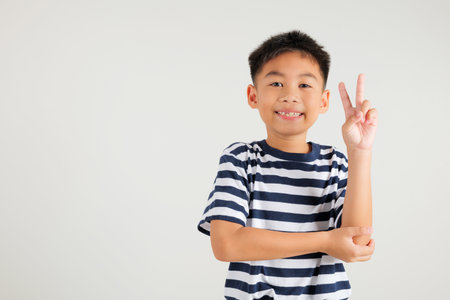 Asian young kid boy showcases optimism and peace by gesturing victory and peace signs in a studio shot with a white background. primary child joyful smile speaks of success, V sign fingerの写真素材