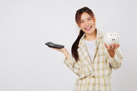 Happy Asian woman compares savings using a calculator while holding a piggybank, then pulls another calculator for tax planning and money accounting. isolated studio white backgroundの写真素材