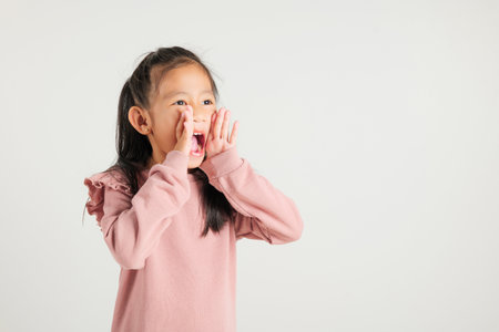 Asian happy portrait cute young kid girl standing hand on mouth talking news announcement studio shot isolated on white background, Thai kindergarten child announcementの写真素材