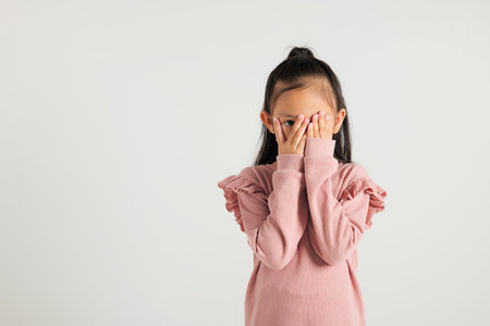 Asian portrait young kid girl in depressed bad mood covering face with hands and peering out with one eye between fingers studio isolated on white background, kindergarten child surprised and shockedの写真素材