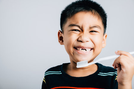 An Asian boy missing an upper milk tooth smiles while holding a toothbrush emphasizing dental care and oral hygiene routine. Isolated on white promoting healthy habits. Children dentist routineの写真素材