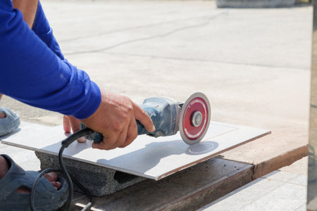 Construction site scene with a worker in blue clothing using an angle grinder to cut a gray tile, highlighting detailed craftsmanship and precision in tile preparation.の写真素材