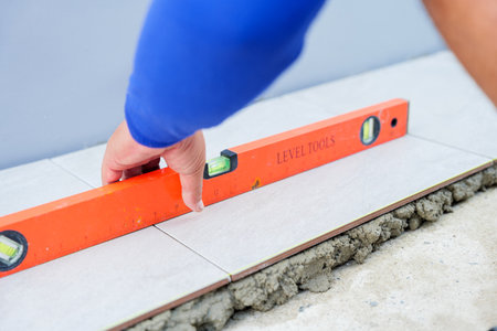 A hand carefully uses an orange level to check the alignment of a tile placed over gray mortar, ensuring precision in tile installation for a flawless finish.の写真素材