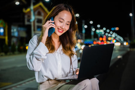 Midst of busy city at night, woman is multitasking on street corner with her laptop and cell phone, utilizing productivity and connectivity of modern technology to stay efficient with urban lifestyleの写真素材