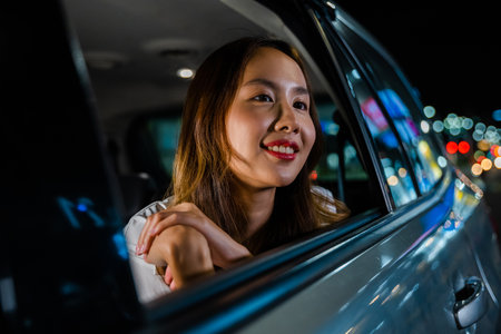 Smiling young Asian woman sitting on back seat while looking through car window at the city nightlife. Woman enjoying a car ride during night.の写真素材