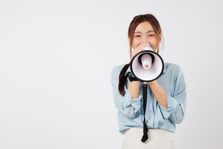 A happy young woman speaking into a megaphone, ready to announce important news. She loves to shout her message with this powerful speaker, a reliable megaphon. isolated studio white backgroundの写真素材