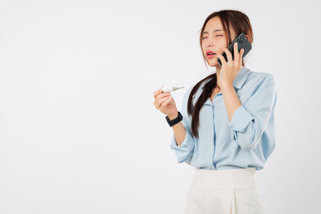 Asian woman feeling worried and sick talks on phone to her doctor about her disease and temperature while checking the thermometer making a call to discuss how sick she is, isolated white backgroundの写真素材