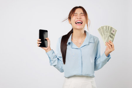 Happy Asian woman showing mobile phone screen and money, excited online app money success, celebrating digital business growth with a confident smile and dollar fan in hand. isolated white backgroundの写真素材