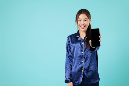 Smiling happy young asian woman in sleep pyjamas is holding mobile phone showing presentation of blank screen workspace on her phone for relaxing at home during bedtime, isolated blue backgroundの写真素材