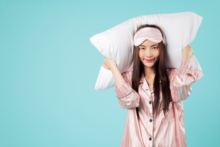 Happy young Asian woman portrait in pajamas holds a pillow on her head, ready for a restful sleep, she is smiling with comfort, wanting more sleep, studio isolated blue backgroundの写真素材