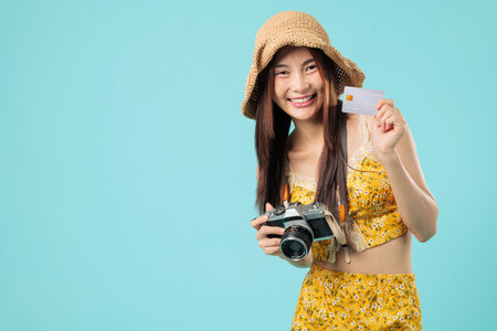 Happy asian woman on travel holds camera, ready to pay online with her bank credit card while wearing a sun hat and floral outfit, holding credit card, headshots isolated studio blue backgroundの写真素材