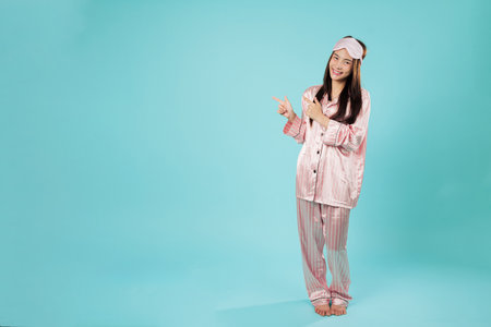 Happy young asian woman in pajamas sleep eye mask rests pointing index finger at blank area, relaxing at home she is calmly pointing left in sleepwear, full body isolated studio blue backgroundの写真素材