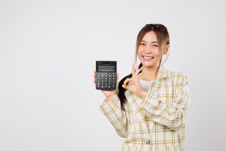 Confident accountant woman presents calculator while holding another calculator, giving OK sign to show marketing approval and business achievement. isolated studio white backgroundの写真素材