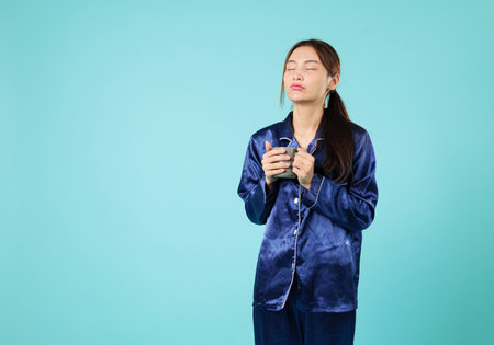 Tired young woman holding a mug of coffee is about to yawn, ready for sleep after a nap, this Asian woman relaxing at home might yawn, looking sleepy, headshots isolated studio blue backgroundの写真素材