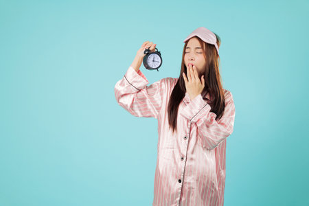 Tired woman yawns holding alarm clock sleep mask, sleep time, pajama and exhausted, checking the time, she looks at the clock with sleepy eyes near clock, headshots isolated studio blue backgroundの写真素材