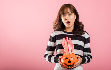 In a Halloween-themed studio shot on pink background, a joyful Asian woman holds model pumpkins, including one in the shape of a ghost. Her funny and festive costume adds to the Halloween spirit.の写真素材