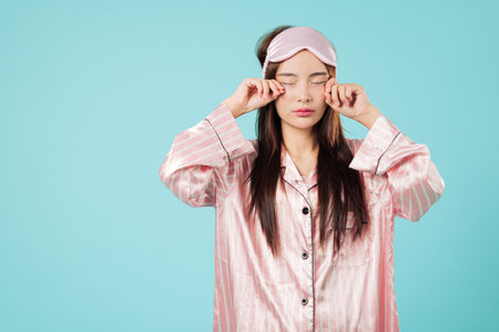 Tired asian woman in pyjamas, sleep mask rubs her eye with fatigue, vision blurred, needs more sleep, wearing pink striped sleep wear and looking sleepy, headshot isolated studio blue backgroundの写真素材