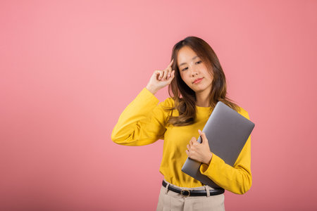 Portrait Asian young woman in a yellow shirt is smiling and holding up her laptop computer and thinking gesture studio shot isolated on pink background, Thai female think her workの写真素材